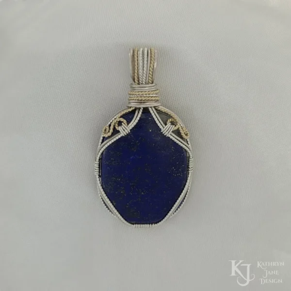 Photo taken in filter cone, eliminating the appearance of reflections off the highly polished surface of the stone Oblong-shaped royal blue lapis lazuli pendant with sterling silver and 14k gold-filled wire spirals at the top, and in the bail. White satin background with Kathryn Jane Design logo in the bottom right