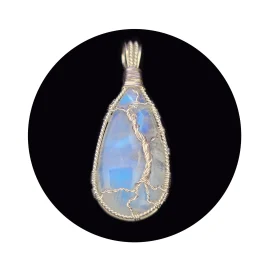 Teardrop-shaped rainbow moonstone sterling silver tree pendant on black background. Blue shimmer reflected back shows effect of partial diamond-shaped halo over a still pool of water in the stone, with the silver wire tree in the foreground, providing support for the stone. The striations on bottom left of the stone look like grasses around the base of the tree, in the foreground of the pool