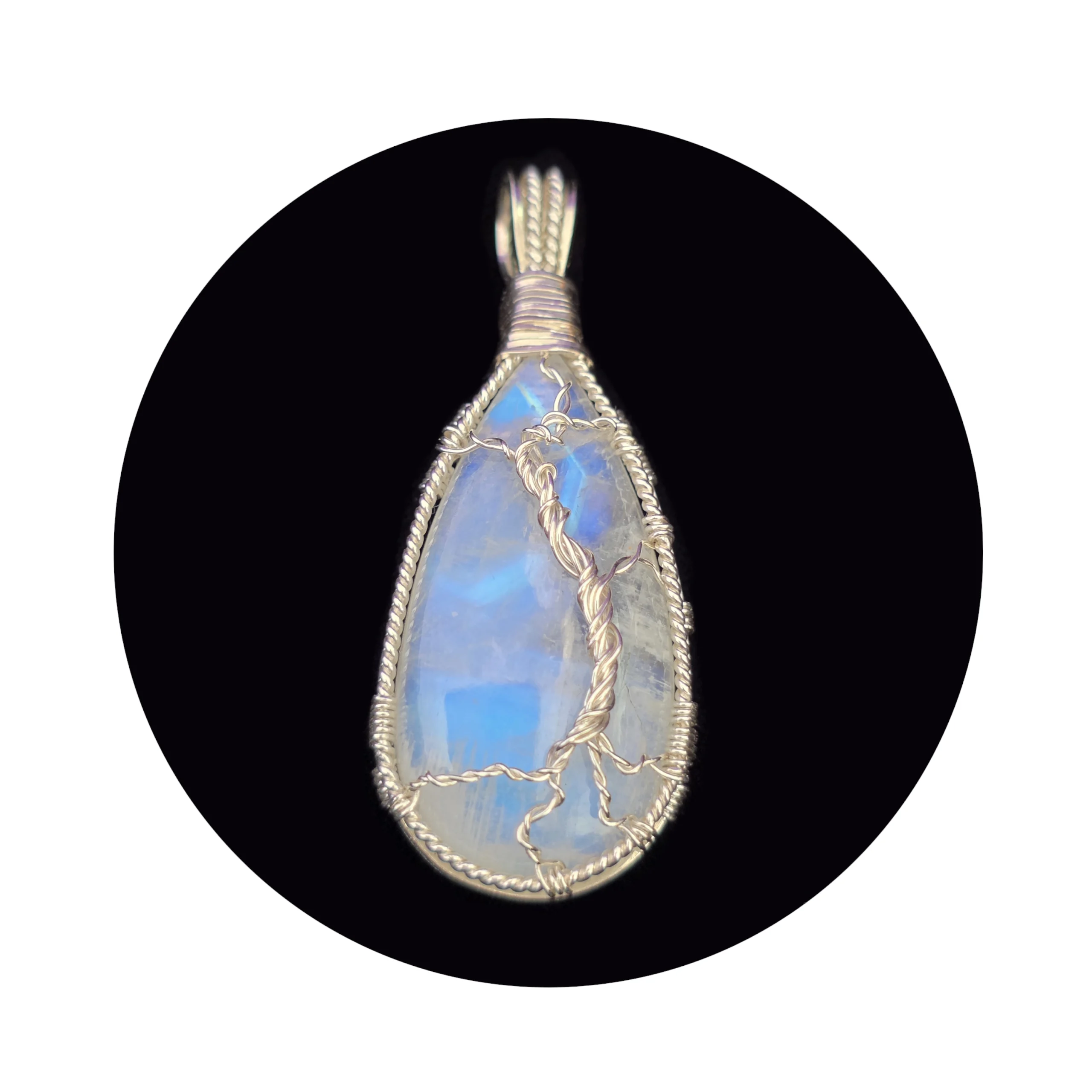 Teardrop-shaped rainbow moonstone sterling silver tree pendant on black background. Blue shimmer reflected back shows effect of partial diamond-shaped halo over a still pool of water in the stone, with the silver wire tree in the foreground, providing support for the stone. The striations on bottom left of the stone look like grasses around the base of the tree, in the foreground of the pool
